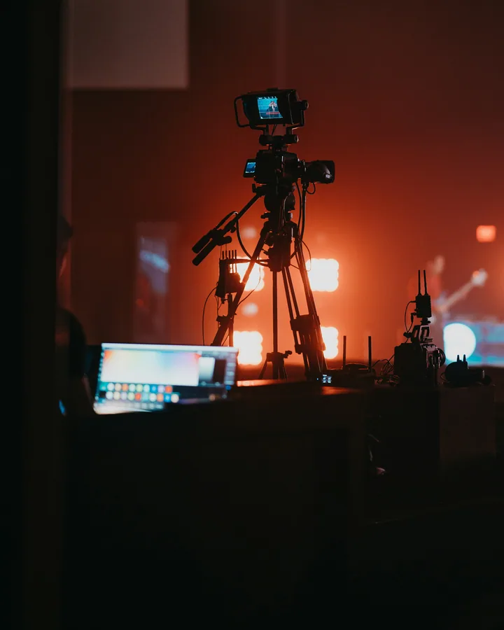 Broadcast camera on a tripod with viewfinder backlit by stage lighting.