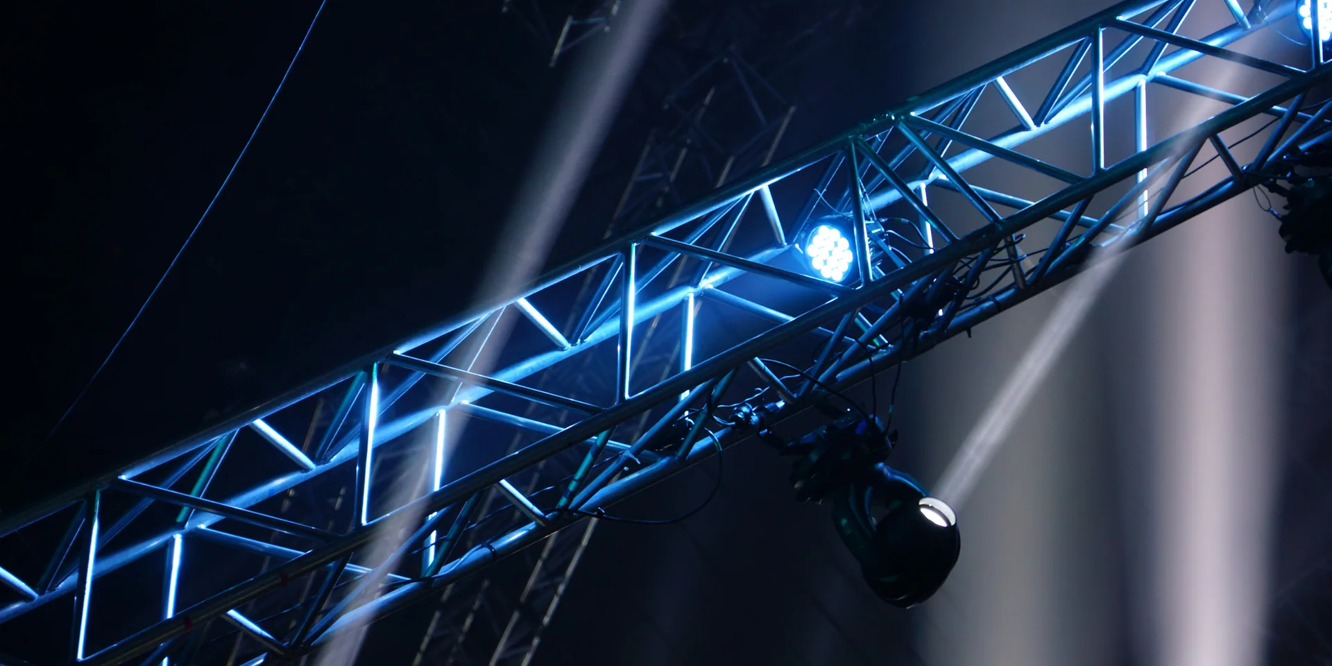 Stage truss with moving-head fixtures and crossing light beams cutting through theatrical haze.