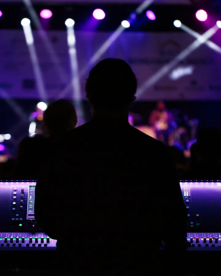 Front-of-house consoles illuminated under stage lights, engineer silhouetted at the desk.