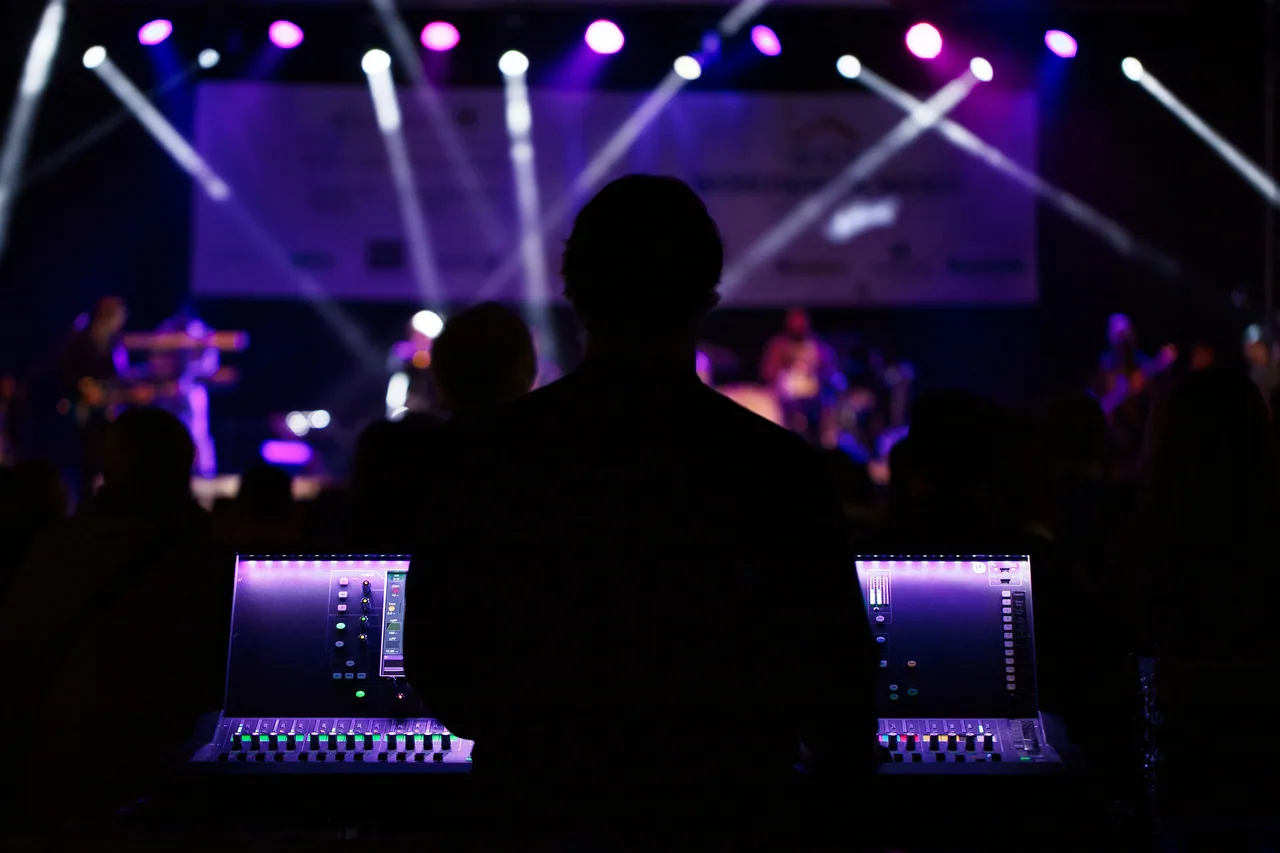 Front-of-house consoles illuminated under stage lights, engineer silhouetted at the desk.