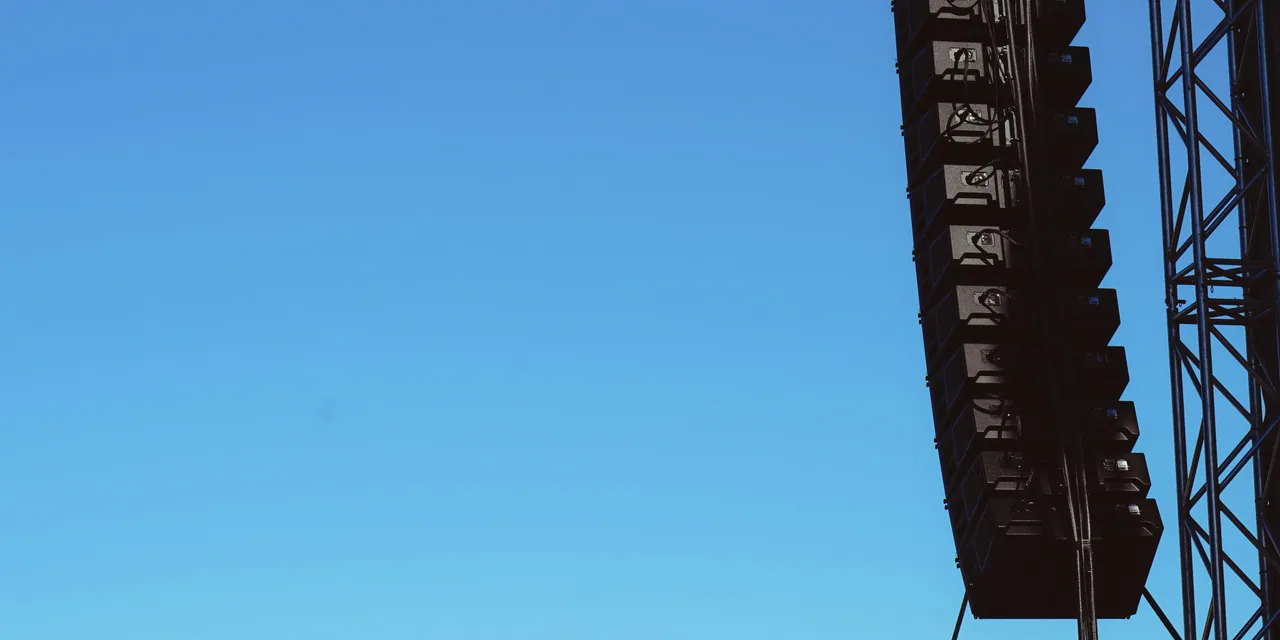 Line-array loudspeakers flown from truss against a clear blue sky.